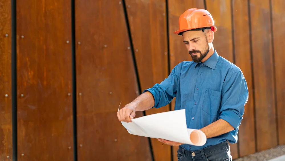 architect.webp An architect in a hard hat reading a rolled drawing in front of a weathering-steel wall